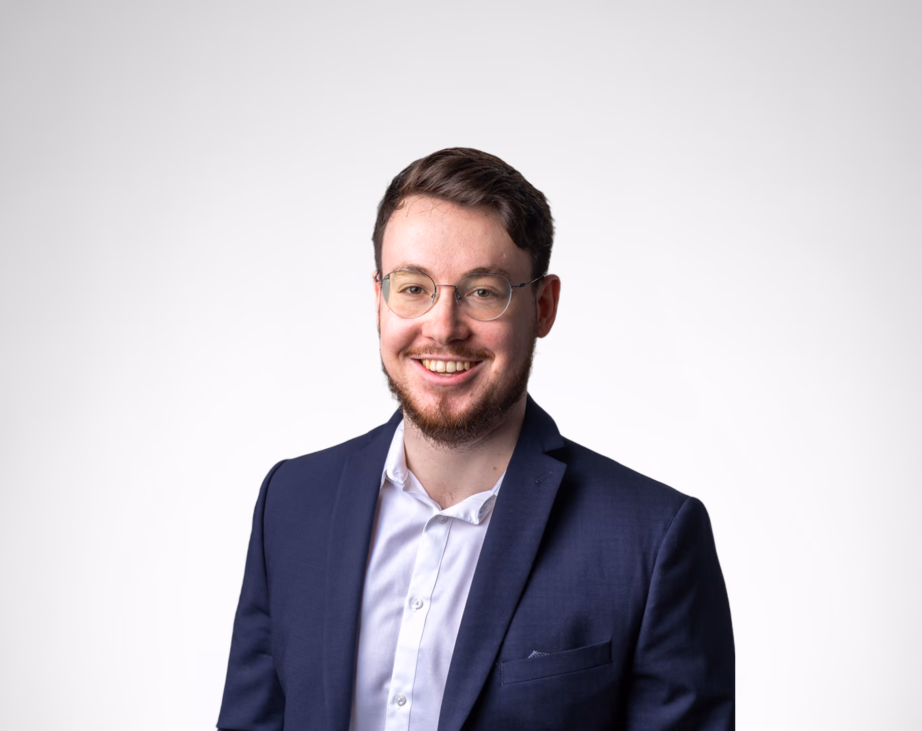 Jeune homme souriant portant des lunettes, une chemise blanche et un blazer bleu marine sur fond clair.