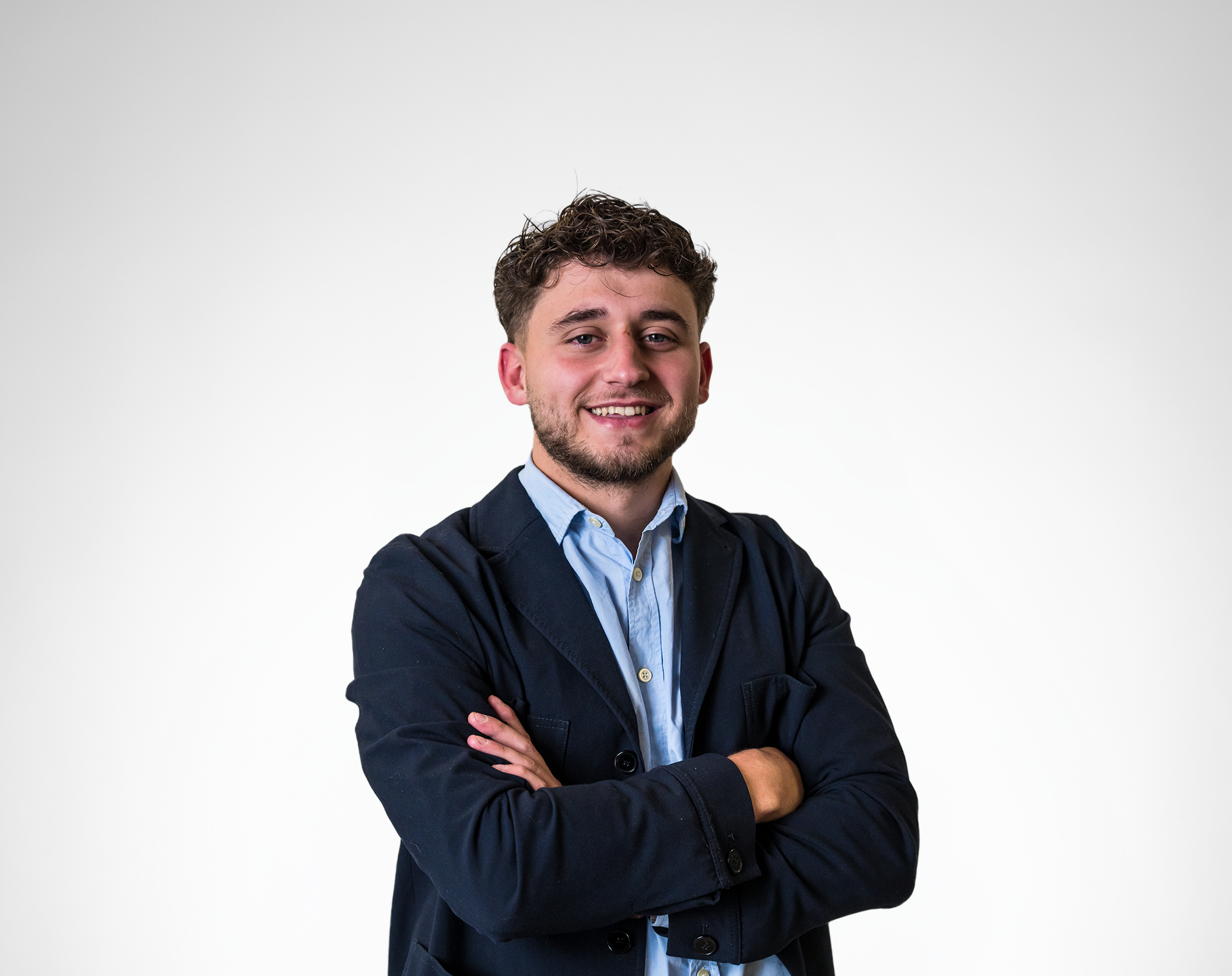 Jeune homme souriant avec cheveux bouclés, barbe légère, portant une chemise bleue et une veste noire, bras croisés devant un fond blanc.