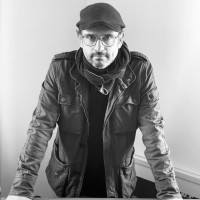 Portrait en noir et blanc d'un homme portant une casquette, des lunettes, et une veste avec un foulard, appuyé sur une table, regardant directement l'objectif.