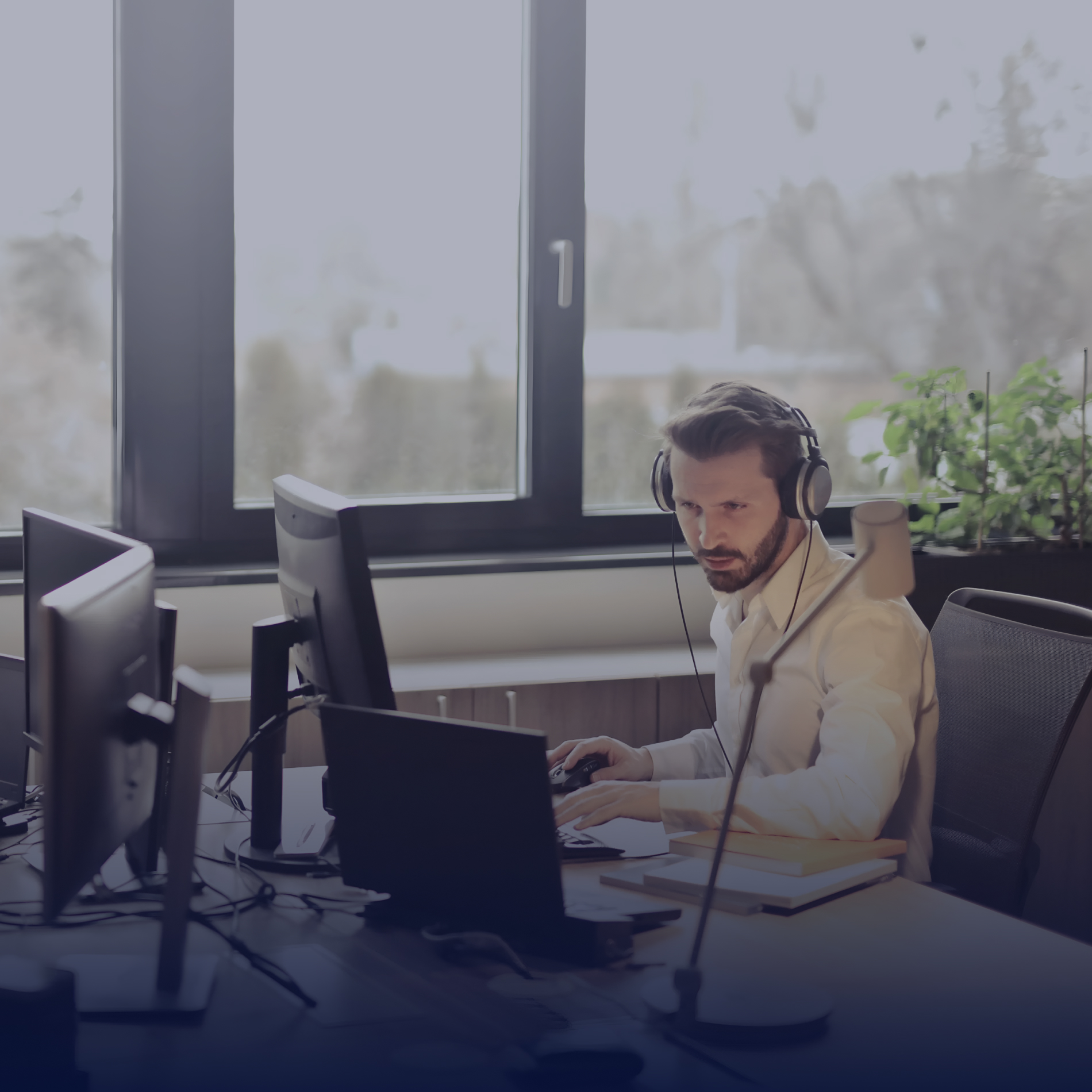 Hombre con auriculares trabajando en un escritorio con dos monitores y una planta al fondo, iluminado por luz natural.