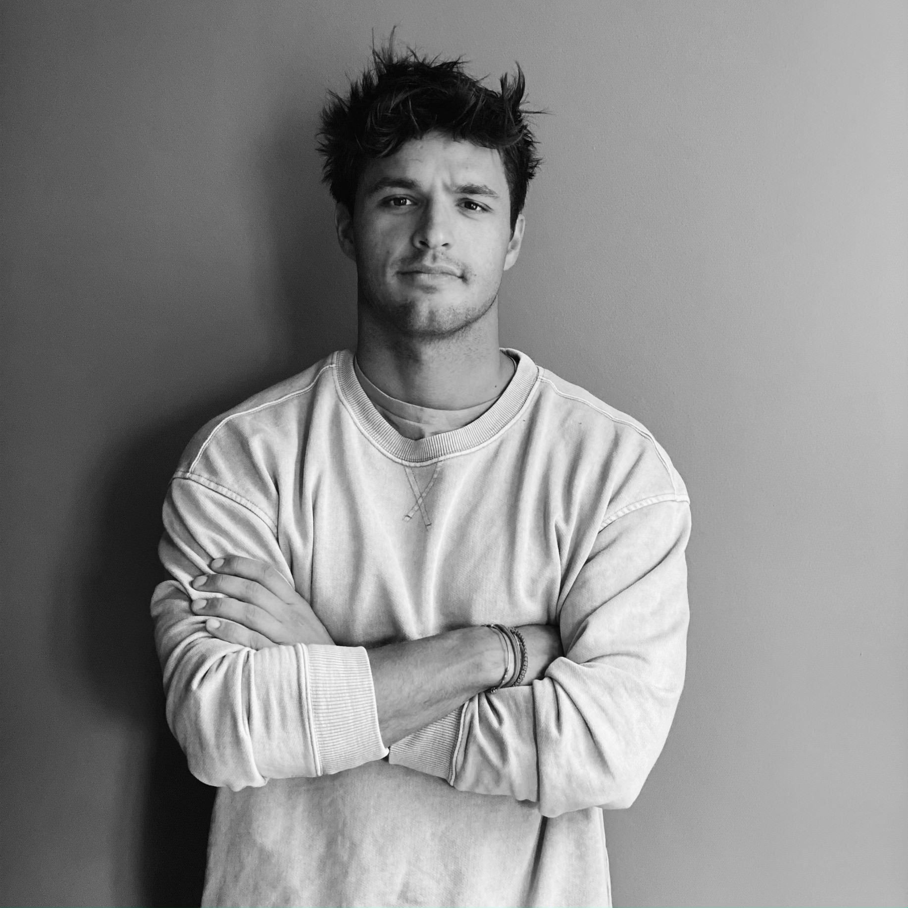 Hombre con sudadera clara, brazos cruzados, frente a una pared gris. Expresión seria y cabello desordenado.
