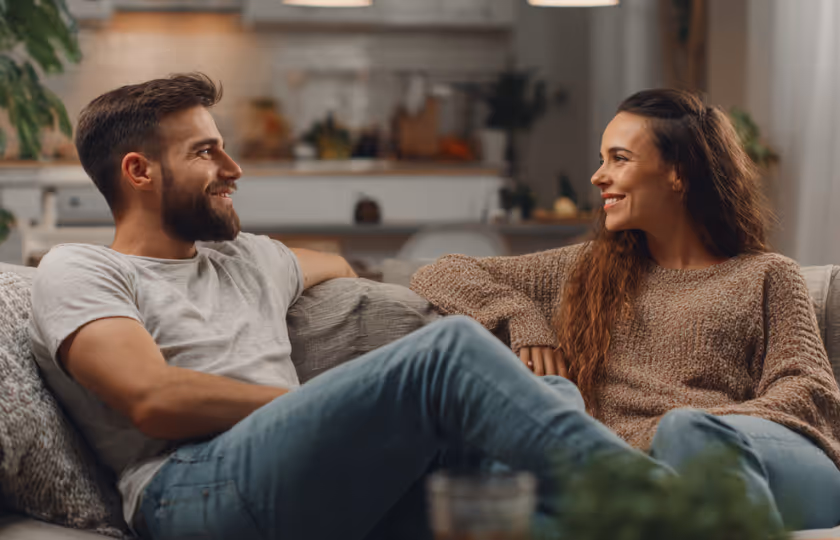 Smiling young man and woman sitting on a couch facing each other in a cozy living room.