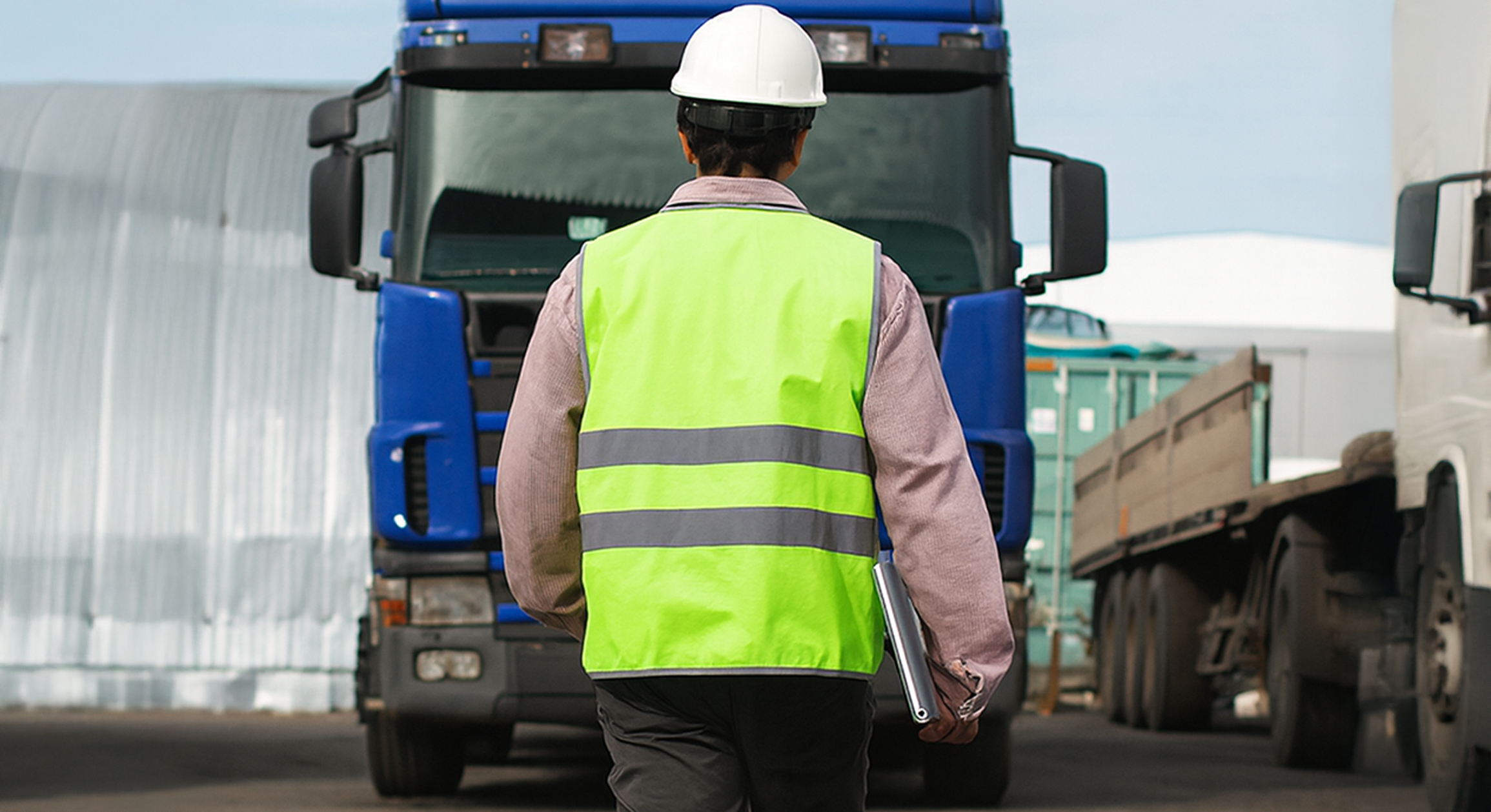 Worker in high-visibility vest and hard hat walking toward a blue freight truck at a logistics yard.