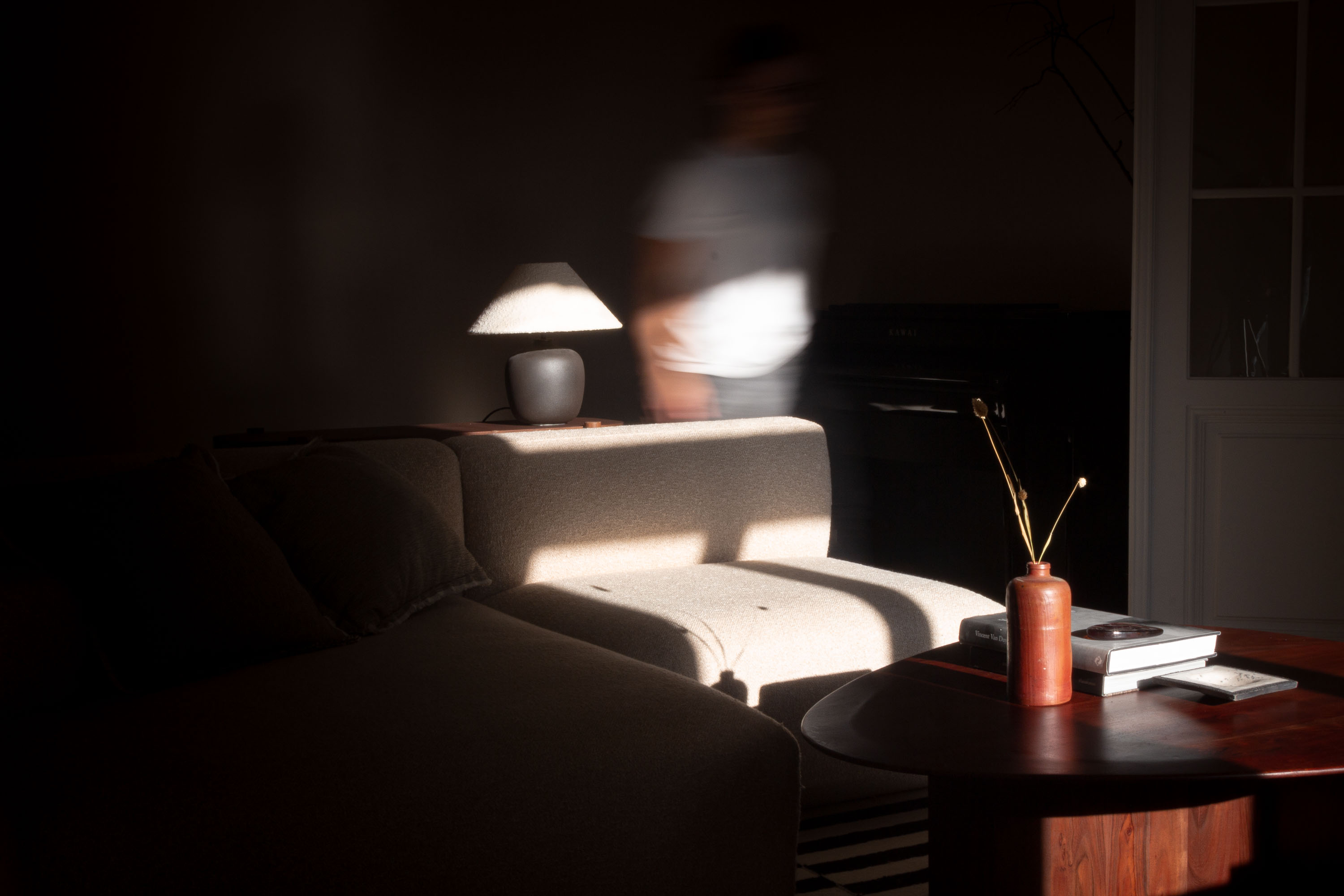 Stimmungsvoller Wohnraum mit Sofa, Couchtisch und Klavier, eine Person in Bewegungsunschärfe, natürliches Tageslicht und dekorative Accessoires.