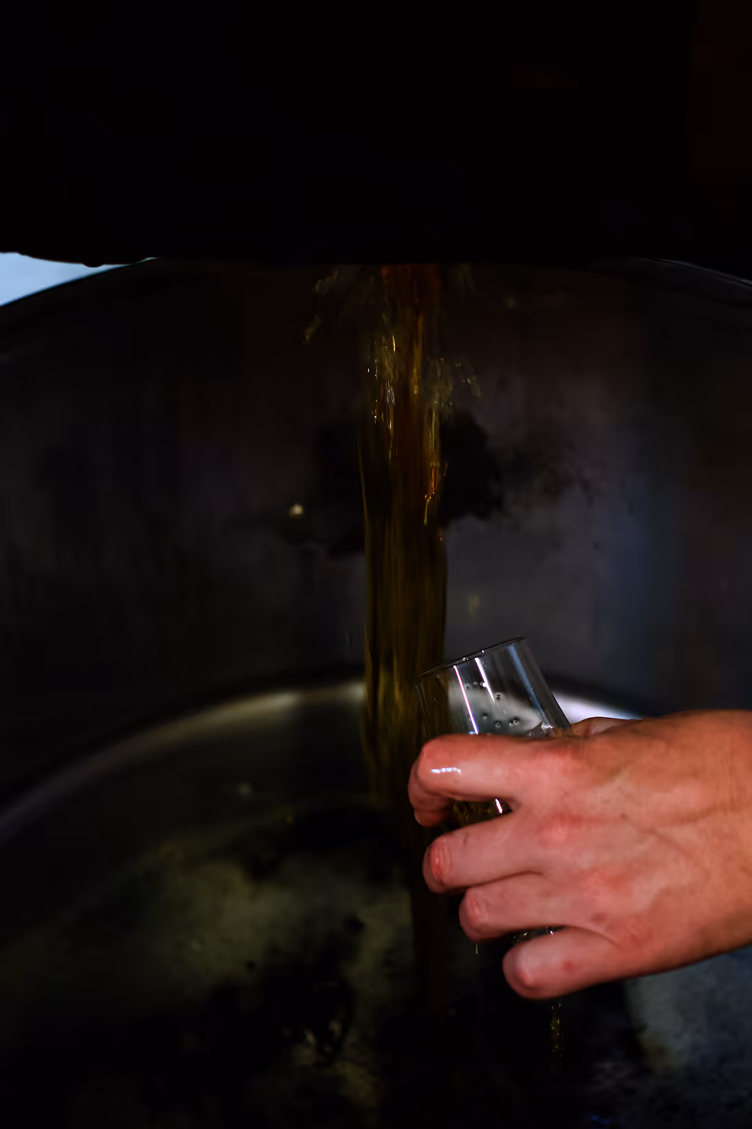 A distiller's hand sampling fresh pot still bourbon whiskey flowing directly from the tank into a glass.
