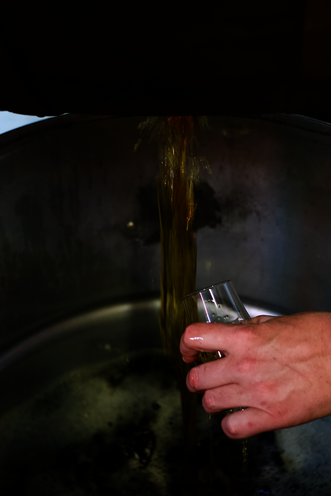 A distiller's hand sampling fresh pot still bourbon whiskey flowing directly from the tank into a glass.