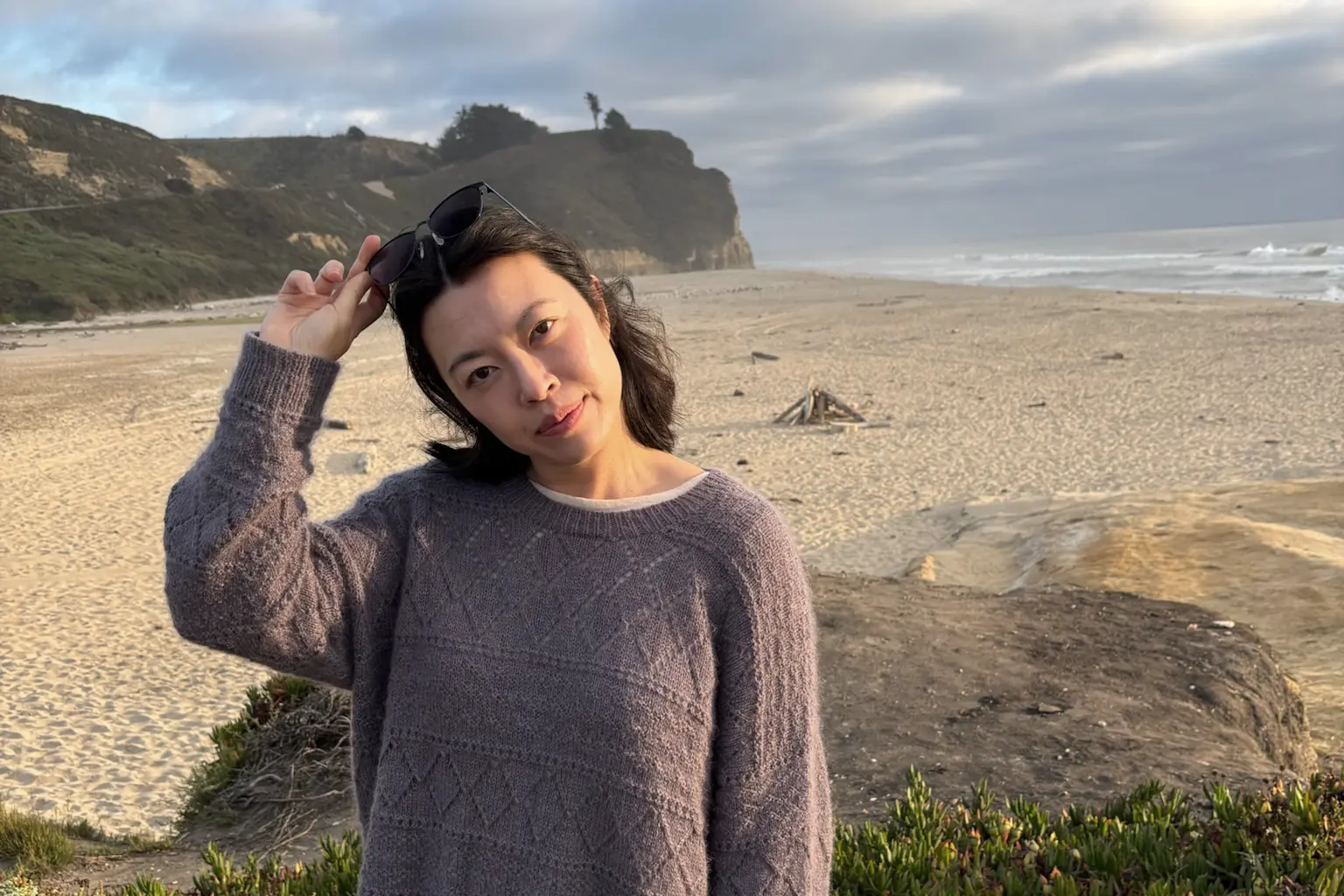 Hanh Le, a Vietnamese woman with shoulder-length dark hair, wearing a purple handknit sweater. She's standing with a beach in the background, at golden hour, adjusting the sunglasses on top of her head.