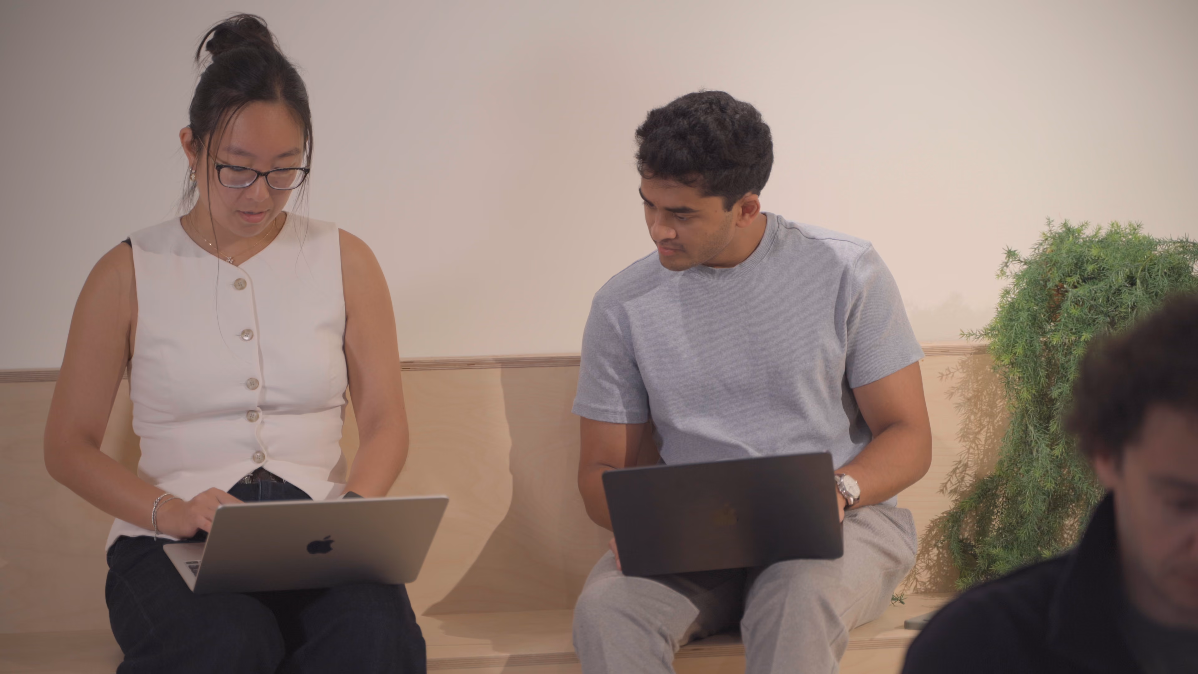A woman and a man sit side by side on a wooden bench, each working on their own laptop, with another person partially visible in the foreground