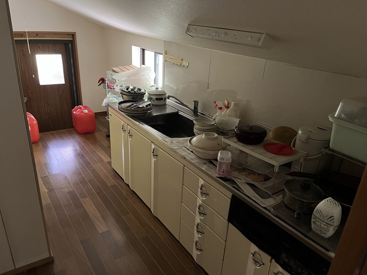 A galley-style kitchen with cream-colored cabinets, dark wooden flooring, and various cooking equipment and dishes arranged on the counter. A door is visible at the end of the hallway.