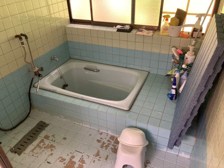 A traditional Japanese bathroom with blue and white tiled walls, a sunken bathtub, and various bath products stored on shelves. The floor shows signs of wear.