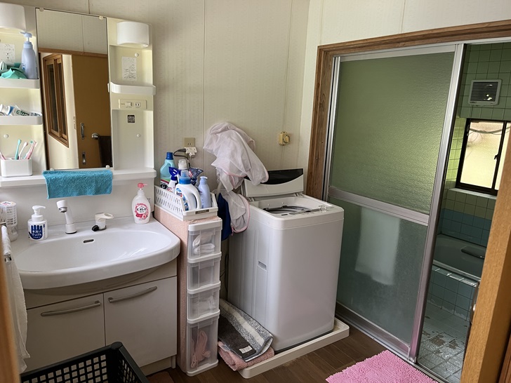 A modern bathroom area with a white sink, washing machine, water dispenser, and various storage solutions. A sliding door leads to what appears to be the bathing area.