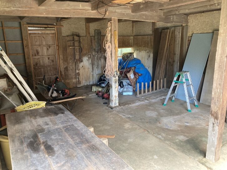 Interior view of a storage or work area with exposed wooden beams, concrete floors, and various tools and equipment stored against the walls. A blue tarp covers some items in the corner.