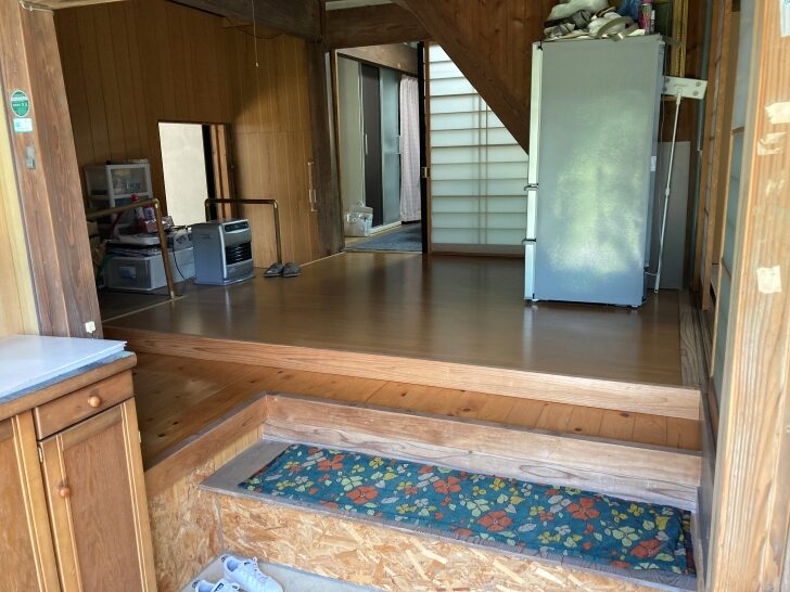 Interior entrance area of a Japanese home showing a traditional step-up floor level change (genkan), with wooden flooring, a decorative floor mat, and household appliances including a refrigerator.