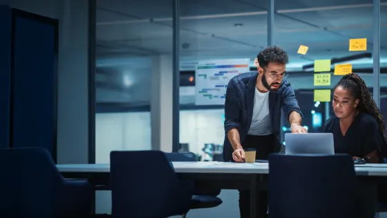 A man and a woman collaborate in a modern office setting, looking at a laptop screen. The man is standing and pointing at the screen, while the woman is seated and looking at the laptop. Sticky notes are visible on a glass partition in the background. The "POWERED BY C7" logo is in the bottom left corner.