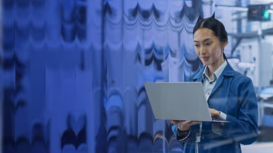 Woman in blue lab coat using laptop in a high-tech industrial or manufacturing facility.