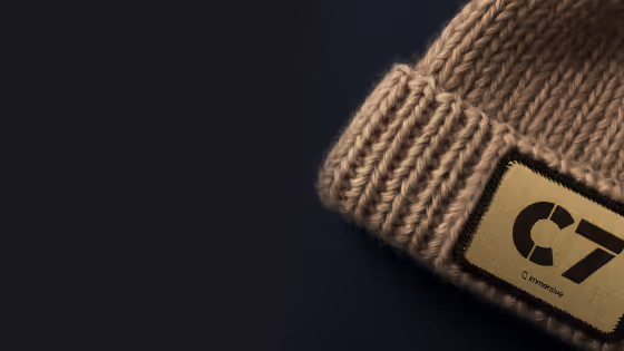 Close-up of a beige knitted beanie with a rectangular C7 patch on a black background.