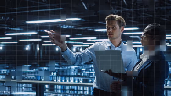 A man points ahead while holding a laptop, speaking to a woman in a dimly lit industrial setting.