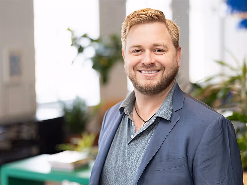Smiling man with beard wearing blue blazer and gray shirt in a bright indoor setting with plants.