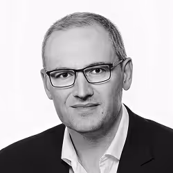 Portrait of a man with glasses wearing a dark blazer and white shirt against a plain background.