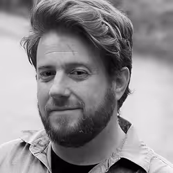 Black and white close-up portrait of a man with a beard and wavy hair, wearing a collared shirt.
