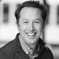 Smiling man with dark hair wearing a collared shirt and jacket in a black and white photo.