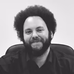 Man with curly afro hair and beard smiling while seated against a plain background.