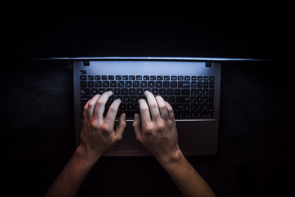 Hands typing on a laptop keyboard illuminated by the screen in a dark environment.