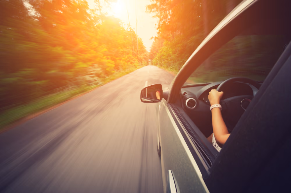 Person driving a car fast on a road surrounded by trees with bright sunlight ahead.