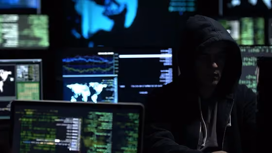 Person in a hooded jacket working with multiple computer screens displaying code and global data maps.
