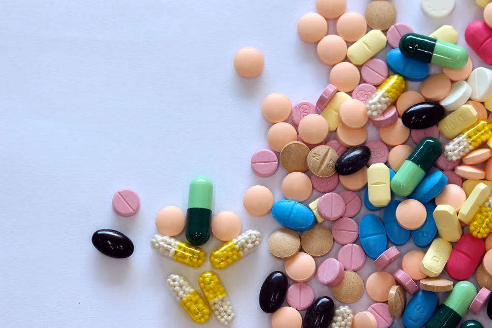 Various colorful pills and capsules scattered on a white surface, including green, blue, yellow, and pink tablets.