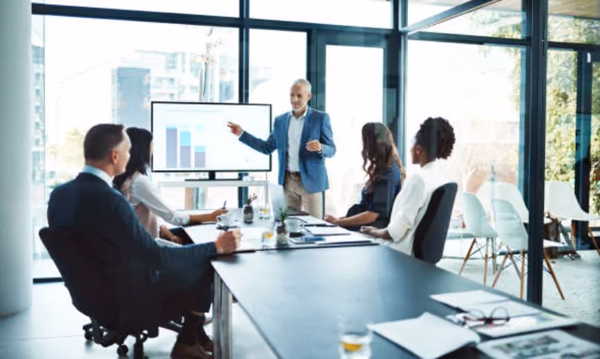 Businessman presenting data on monitor to colleagues seated around conference table in modern office.