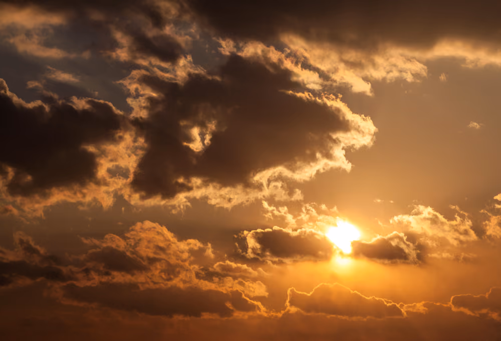 Sun glowing through dark clouds in an orange sky at sunset or sunrise.