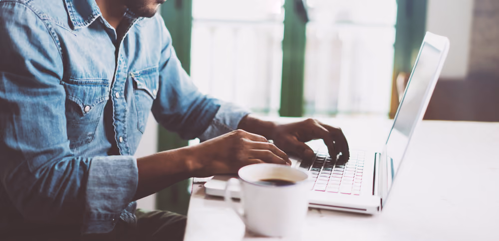 Person wearing denim shirt typing on a laptop with a cup of coffee on the table.