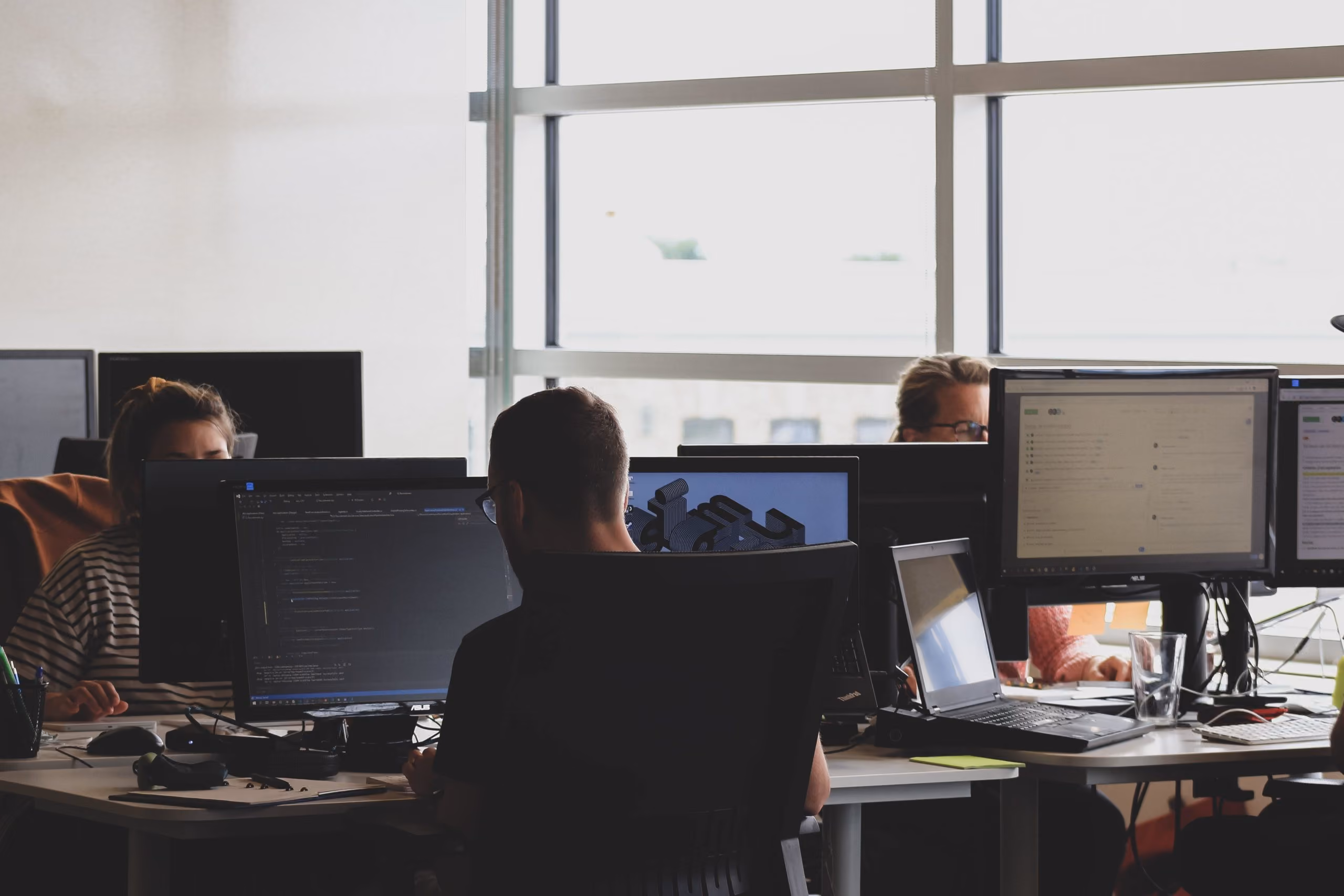 People working on computers in an office with large windows letting in daylight.