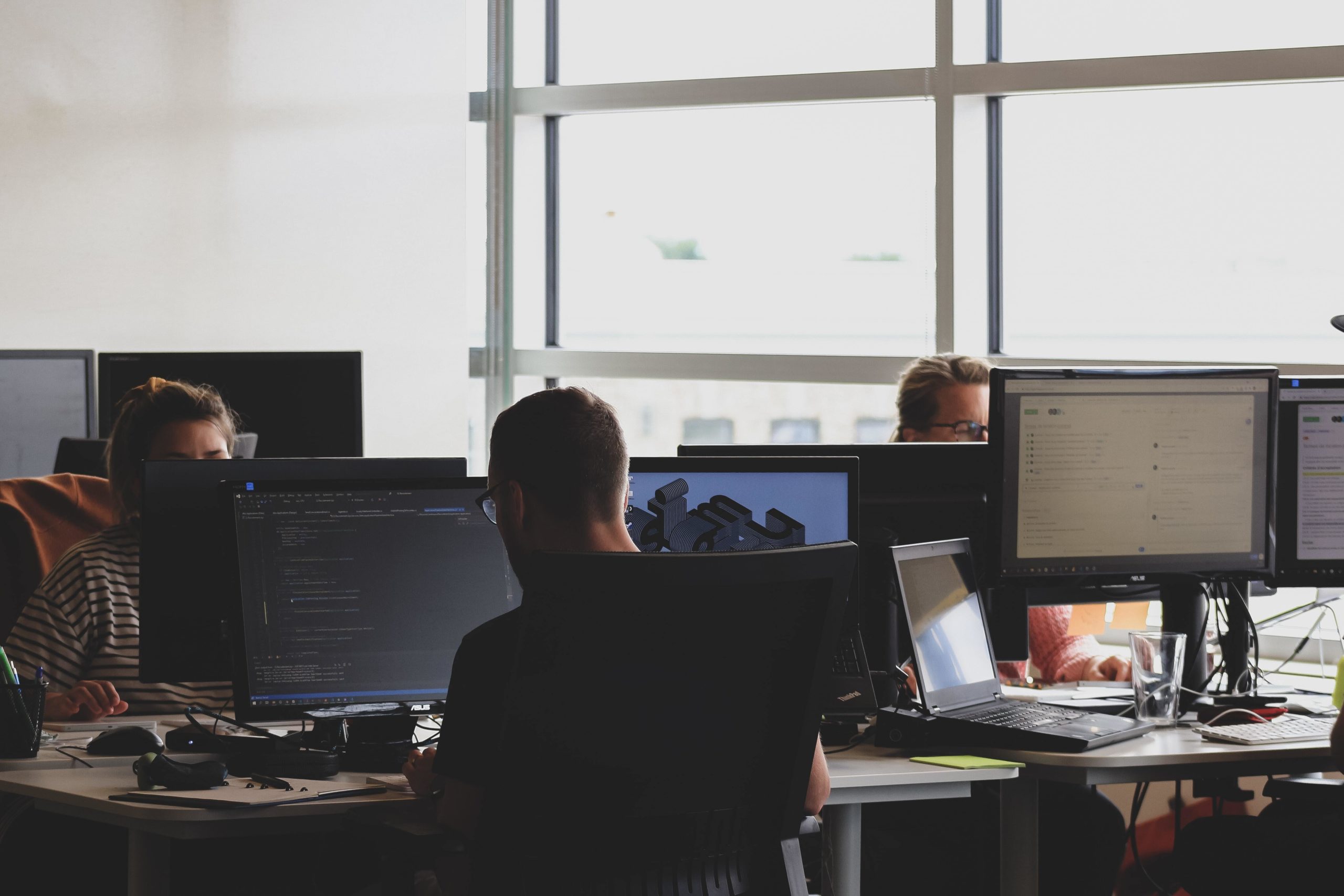 People working on computers in an office with large windows letting in daylight.