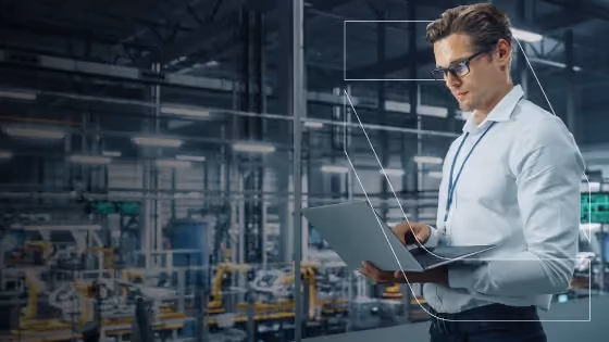 Man wearing glasses and ID badge using laptop in a modern industrial factory setting.