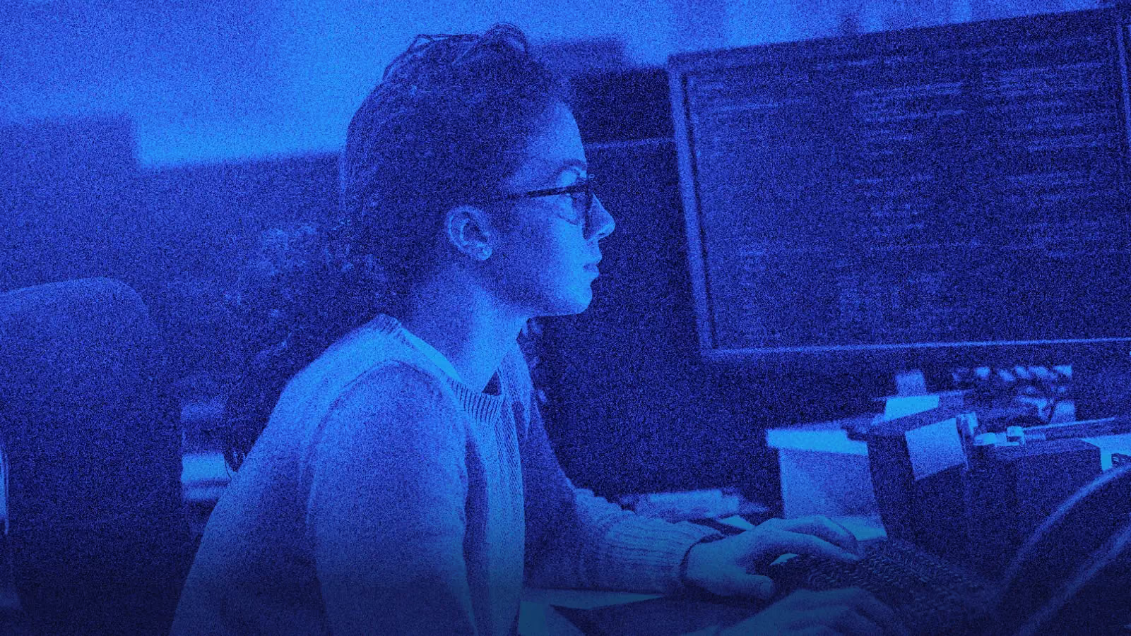 Woman with glasses focused on coding at a computer in a dimly lit, blue-toned room.