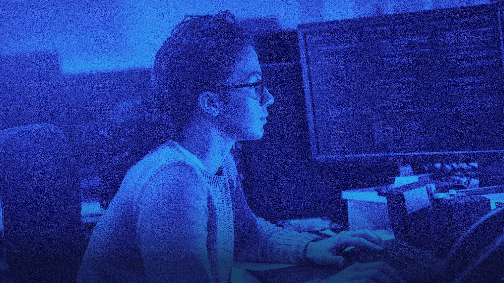 Woman with glasses focused on coding at a computer in a dimly lit, blue-toned room.