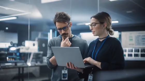 Two office workers wearing ID badges reviewing content on a laptop in a modern workspace.