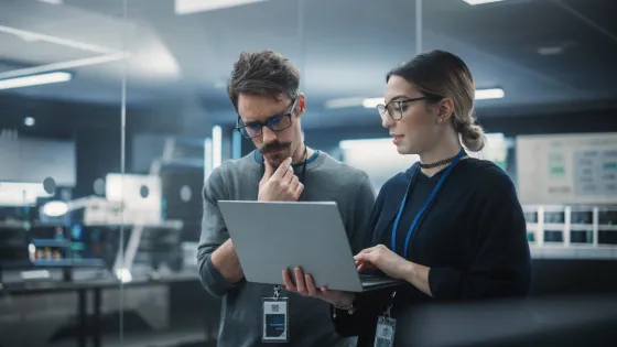 Two office workers wearing ID badges reviewing content on a laptop in a modern workspace.