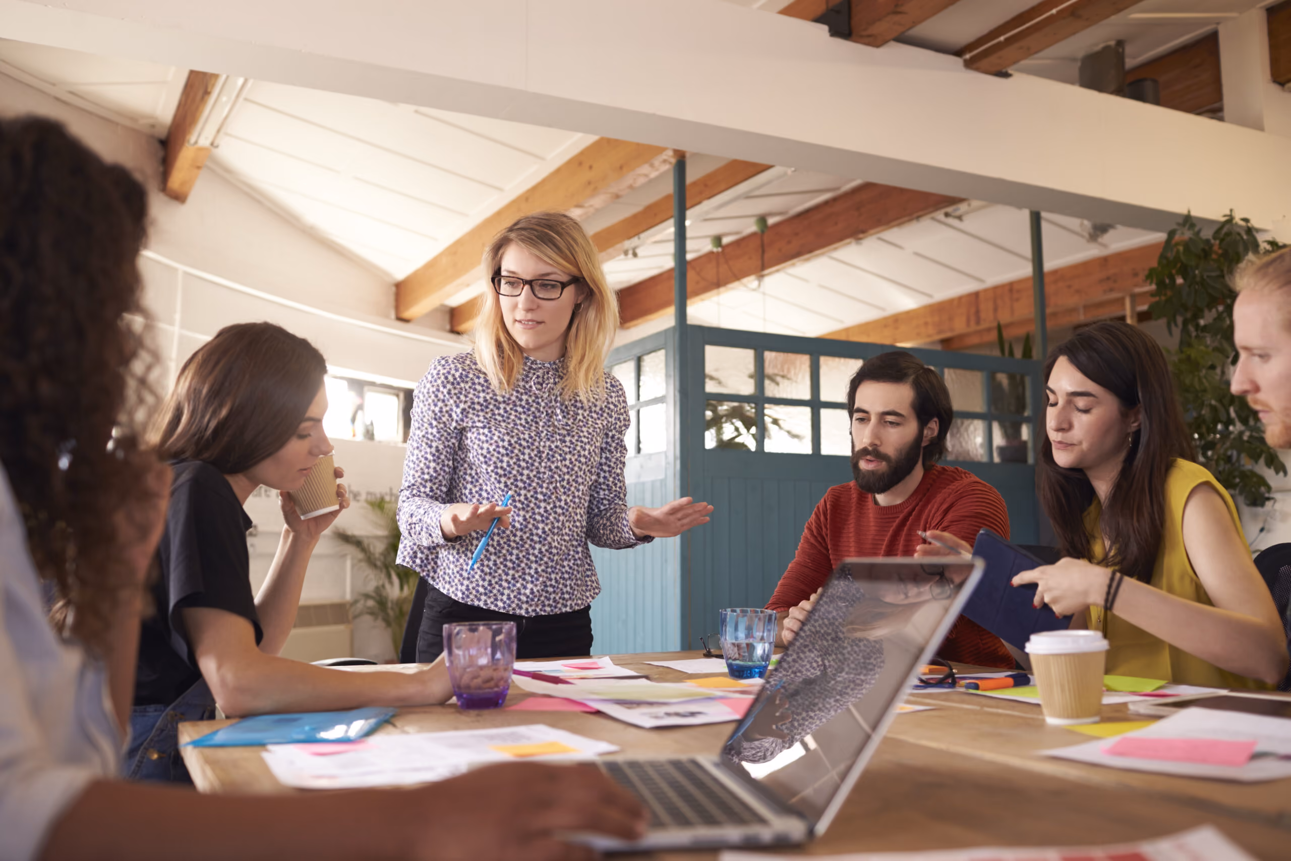 Five diverse coworkers discuss ideas around a table with laptops, papers, and coffee in a bright office.