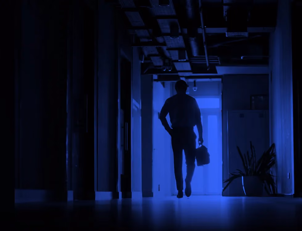 Silhouette of a person walking down a dimly lit hallway carrying a briefcase or bag.