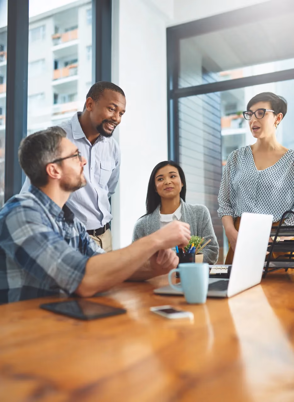 Four diverse coworkers discussing work around a table with laptop, phone, and mug in a bright office.