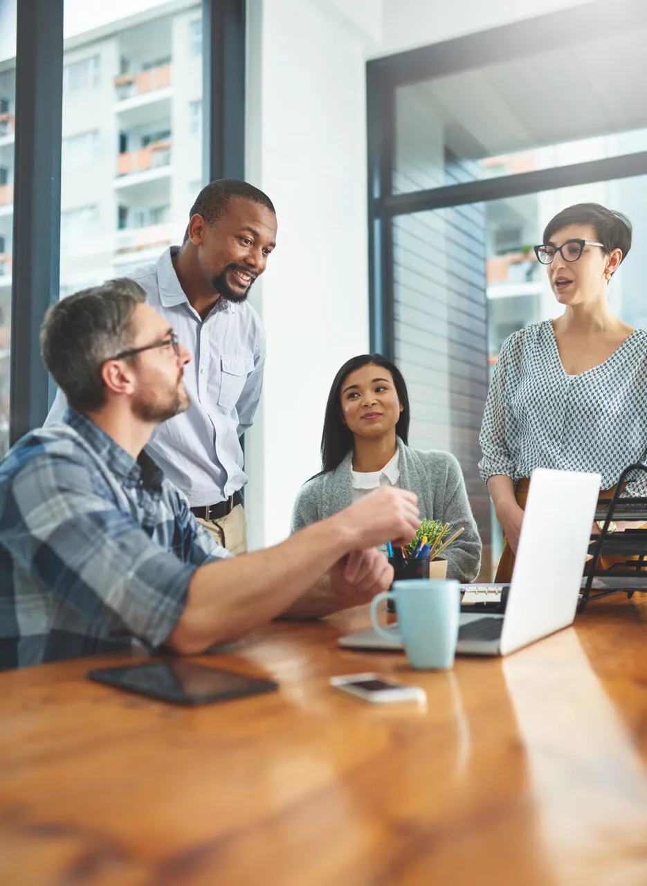Four diverse coworkers discussing work around a table with laptop, phone, and mug in a bright office.