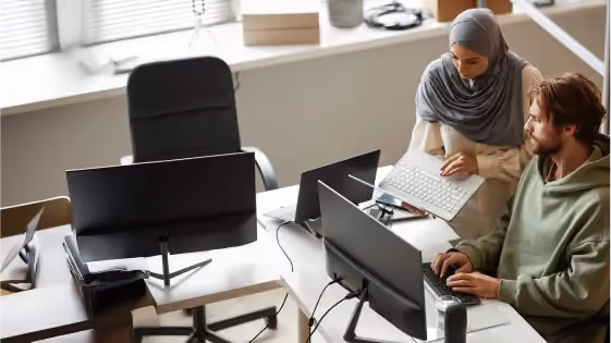 A man and woman collaborating at a desk with multiple computer monitors in a bright office.