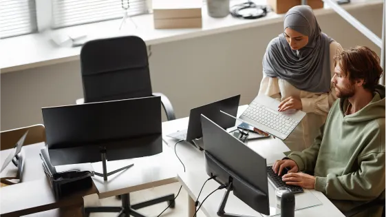 A man and woman collaborating at a desk with multiple computer monitors in a bright office.