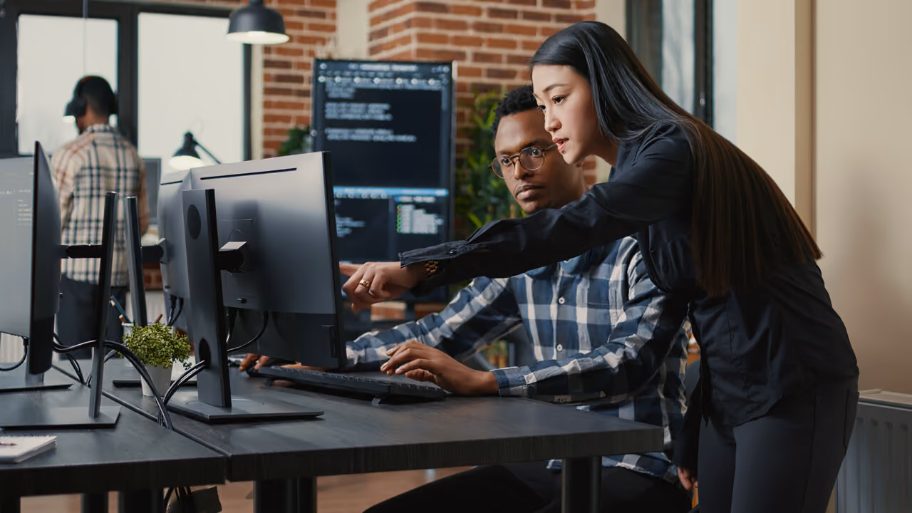 Woman pointing at a desktop computer screen while a man in glasses looks on attentively in an office.