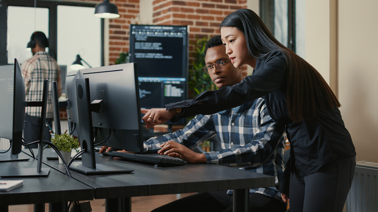 Woman pointing at a desktop computer screen while a man in glasses looks on attentively in an office.