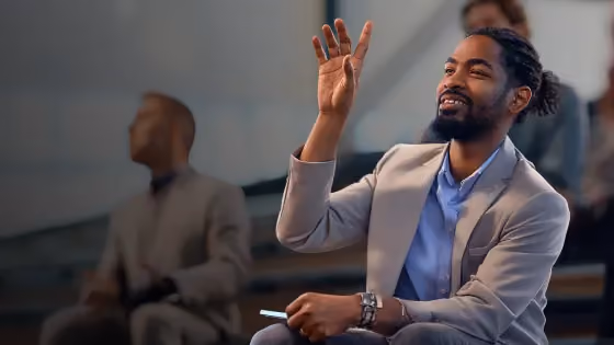 Man in a suit raising hand, holding smartphone, with another person blurred in the background.