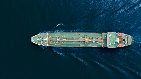 Aerial view of a large green cargo ship sailing on dark blue water leaving a wake behind.